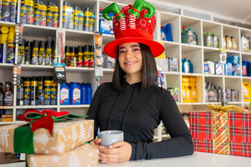 Woman wearing holiday elf hat and holding a mug, standing near christmas gifts
