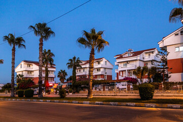 Quiet residential street with white three-story houses and tile roofs under post-sunset blue hour, palm trees lining the road in typical Turkish Mediterranean neighborhood. Side, Antalya, Turkey.