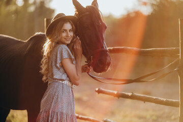 A young woman with a horse stands near a fence in autumn at sunset. Breeding thoroughbred horses for equestrian sport.