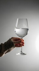 A hand wearing a textured sweater holds up a large clear glass filled with sparkling water and ice cubes against a soft gray background