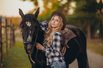 Obraz premium A young woman with a horse stands near a fence in autumn at sunset. Breeding thoroughbred horses for equestrian sport.