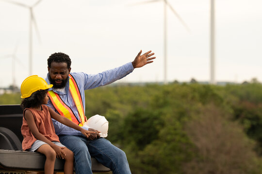 African American engineer father pointing at wind turbines while sitting with daughter, symbolizing guidance, renewable energy education, family connection, and inspiring future in clean technology.