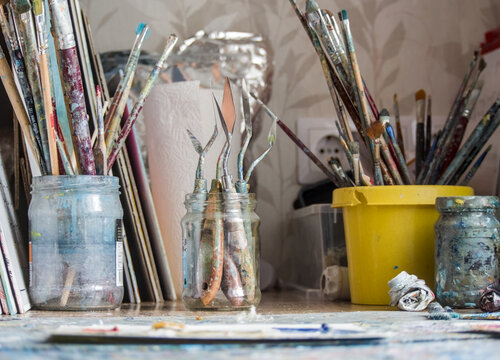 Artist's paint brushes - A messy artist's desk with numerous paintbrushes of all sizes in glass jars and buckets - Powered by Adobe