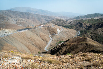 A winding mountain road cuts through the dry slopes of the High Atlas in Morocco, offering a breathtaking view of rugged peaks under a hazy blue sky.