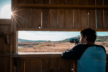 Man with camera photographing nature through a wooden bird hide window. Warm sunlight and dry...