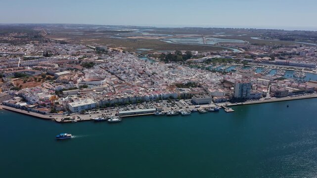 Aerial view drone shot of Ayamonte Aerial view bay approach with passenger ferry crossing calm turquoise water toward sandy headland, seaside town backdrop and distant marshlands.