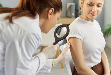 Female doctor examining hand skin of young woman patient. Professional dermatologist using magnifying glass checking skin condition in clinic. Dermatology, skin health care concept.