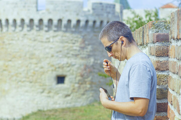 A man puts on wired earbuds connected to a smartphone while standing against castle towers. Symbolizes personal connection across distance, communication in travel, and digital nomad lifestyle.