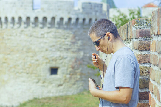 A man puts on wired earbuds connected to a smartphone while standing against castle towers. Symbolizes personal connection across distance, communication in travel, and digital nomad lifestyle.