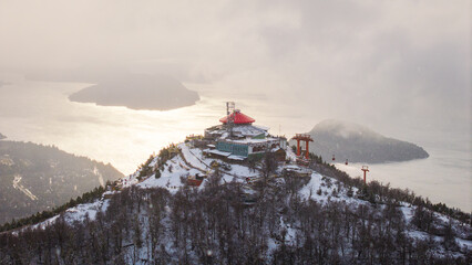 Aerial winter view of Cerro Otto with red mountain refuge and Nahuel Huapi Lake reflecting sunlight in Bariloche, Argentina.