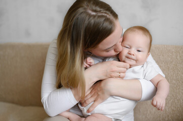 Mother holds sweet baby while sitting on couch, kissing his chubby cheek. Tender maternal hug. Mother with newborn at home. Infant care concept.