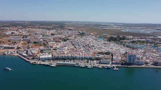 Aerial view drone shot of Ayamonte Aerial view coastal town and ferry approaching bay, whitewashed buildings lining seafront, marina mouth and long ocean horizon under clear sky.