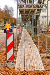 Temporary pedestrian walkway under metal scaffolding at city construction site. Safety barrier, warning light, and autumn leaves on the ground.