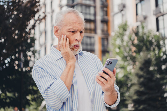 Elderly man in casual striped shirt checks smartphone outdoors in the city with modern buildings in the background