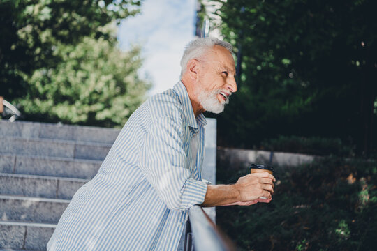Older man with gray beard smiles while leaning on a railing and holding a coffee cup outdoors in a sunny city park near modern buildings and urban scenery