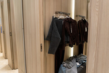 Several garments are hanging and folded inside a fitting room with light wood panel doors. The...
