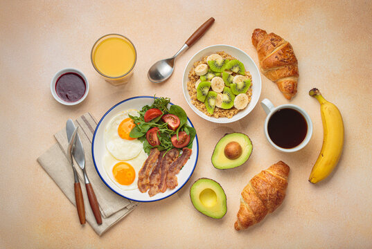 Breakfast table healthy foods fried eggs, bacon, oats fruit, avocado, croissants, coffee, orange juice warm beige background top view