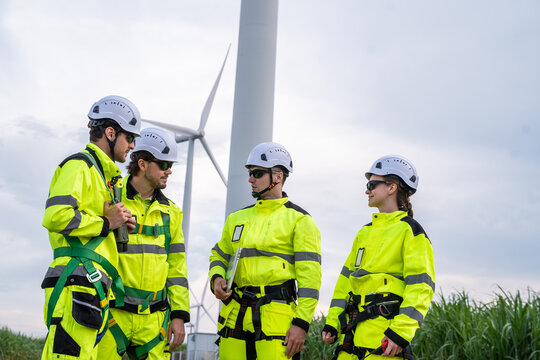 Four people in yellow and white safety gear are standing near a wind turbine. They are wearing helmets. Professional technicians with PPE and harnesses preparing for maintenance at wind farm. - Powered by Adobe