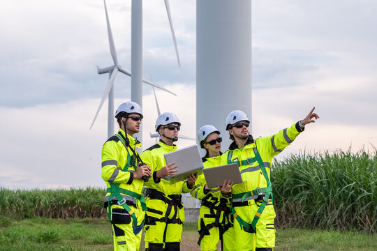 A group of four workers in yellow safety gear are standing in front of a large wind turbine. Professional technicians with PPE and harnesses preparing for maintenance at wind farm.