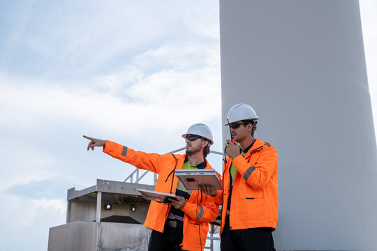 Two men in orange jackets are pointing at a building. One of them is holding a clipboard. They are wearing hard hats. Technicians team collaborating on renewable energy project at wind farm site. - Powered by Adobe