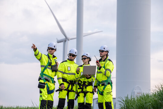 A group of four workers in bright yellow and black safety gear are standing near a large wind turbine. Professional technicians with PPE and harnesses preparing for maintenance at wind farm.