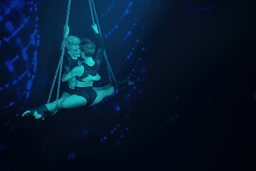 Two female aerialists on chains under cool teal light—performing a midair split and embracing in a tender moment just before a kiss; horizontal frame with wide right-side negative space for copy.
