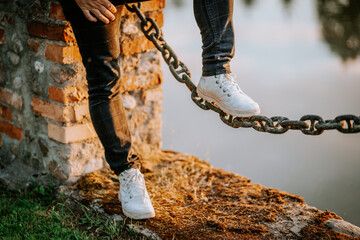 Individual balancing on a large chain over water, wearing white sneakers and dark jeans, showcasing a moment of daring and adventure in a serene outdoor setting