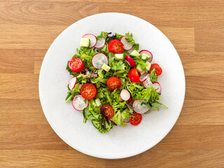 Simple salad with lettuce, tomatoes, cucumber and radish on a white plate on a wooden table.