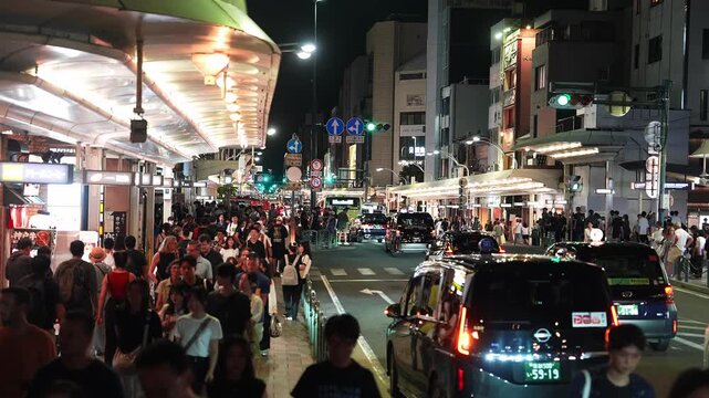 Night street in downtown Kyoto shows crowds under a curved lit canopy as taxis, a city bus, and cars move slowly. Blue traffic signs and lamps define the scene.