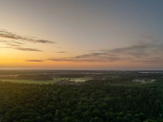 Naklejka premium Aerial drone view of Vinni, Estonia, at sunrise. Golden sky meets soft morning mist over lush green forest and open fields. Tranquil summer landscape with warm pastel tones and calm early dawn.