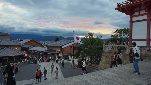 Dusk view from Kiyomizu dera stone plaza in Kyoto, Japan. Tourists move on steps, flag and komainu stand midground, tiled roofs lead to city and mountain silhouettes.