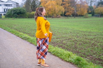 Happy Mature woman in mustard sweater and plaid skirt walking outdoors, holding yellow bag, autumn style, confident mood, in bright fall outfit walking on path, enjoying life surrounded by lush nature
