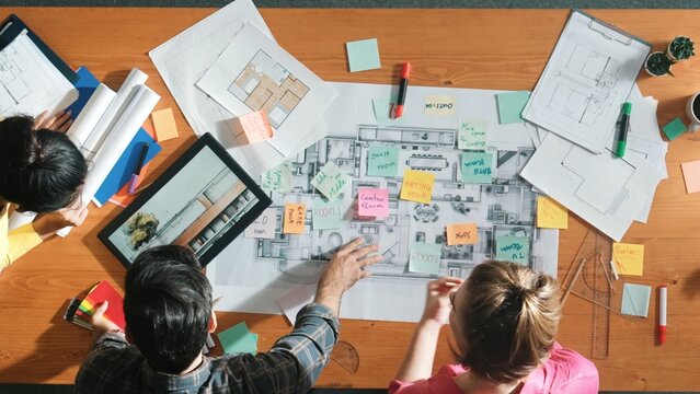 Top down aerial view of interior designer team choosing color from palettes while architect engineer planning house construction from tablet and project plan at table with building plan. Symposium.