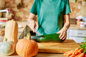 A woman in the kitchen in a green T-shirt cuts zucchini on a wooden board with a knife. Ripe orange pumpkins on a wooden kitchen table, space for a copy. Thanksgiving Dinner Preparation