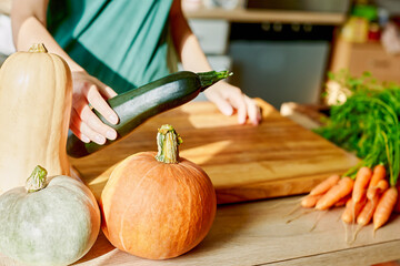 A woman's hand with a large green zucchini in the kitchen is cooking. Pumpkins ripe on kitchen wooden table, space for copy. Udin for Thanksgiving, Halloween, Fall Holiday