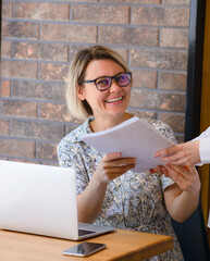 A young laughing woman is handed papers in her hands, an open laptop is on the table. A businesswoman works at a wooden table with a laptop, a girl hands over documents to her boss, a teacher takes a