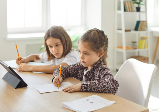 Two children sitting at desk in room focused on writing with pencils and doing homework. Siblings concentrating on learning activities, studying or drawing, using digital tablet at home together.
