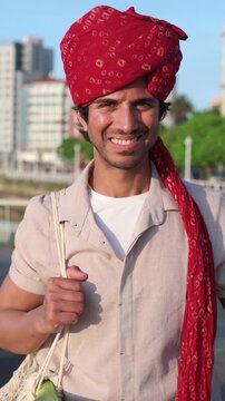 Portrait of a handsome young indian man wearing a traditional red turban, looking at the camera and smiling, standing outdoors in a city