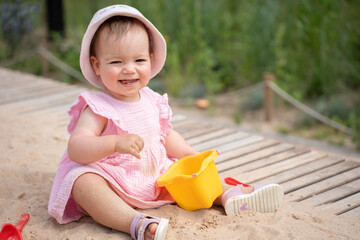 adorable toddler in pink dress and sun hat playing joyfully with a yellow bucket in a sunny park setting, sitting on sandy pathway with soft greenery in the background, relaxed, moment, innocence
