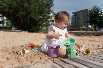 child in floral dress engaged in playful sand activities with colorful toys in a sunlit urban park on a bright day with greenery and buildings in the background, peaceful, relaxation, childhood