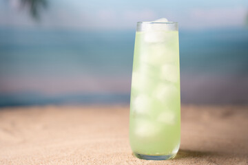 chilled green beverage with ice in clear glass on sandy beach with blurred ocean backdrop creates refreshing tropical vibe perfect for relaxation and summer enjoyment, seaside, escape, rejuvenating