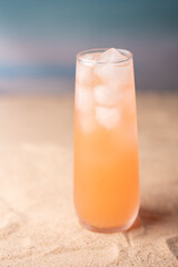 chilled peach-orange beverage in tall glass with ice cubes on sandy beach setting against blurred blue and sandy backdrop evoking refreshing summertime vibes, contentment, repose, placid, inviting