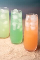 refreshing trio of iced beverages in tall glasses on sandy beach setting with vibrant orange and green hues against a tranquil blue backdrop, invoking a cool, summery atmosphere, respite, warm