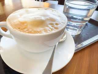 Warm cappuccino with foam and glass of water on tray in sunlit cafe