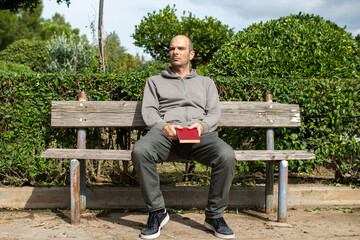 Fototapeta premium Photo of a man sitting on a bench alone holding a red book at his hands.