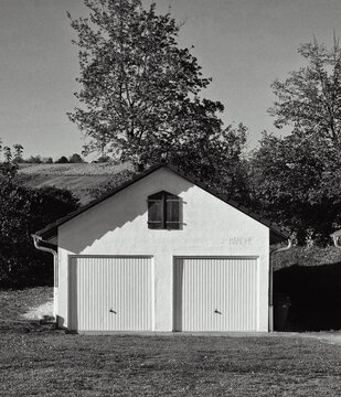 Symmetrische Schwarzwei&szlig;-Aufnahme einer einfachen Doppelgarage oder eines Nebengeb&auml;udes mit Satteldach. Zwei Holztore und ein kleines Fenster mit Fensterl&auml;den, umgeben von B&auml;umen und einer sanften H&uuml;g