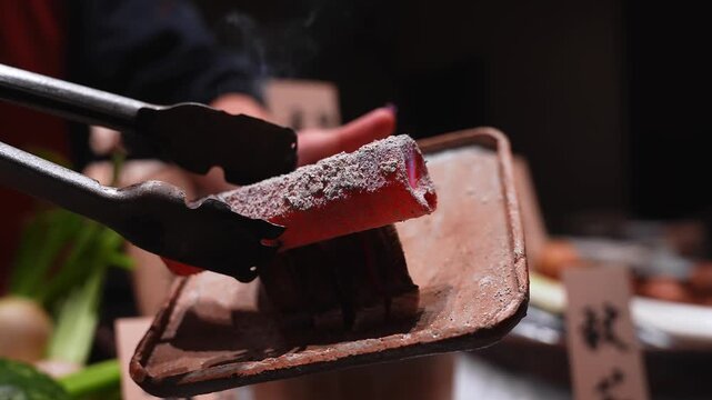 Metal tongs hold a glowing ember above a clay grill as food sears at a Tokyo stall. A vendor adjusts charcoal, smoke rises, wooden labels blur in warm night light.