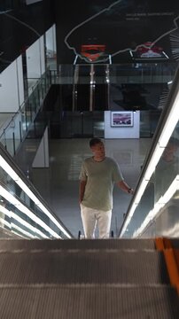 A casually dressed man rides an illuminated glass escalator in a multi level automotive exhibit with a 24h Le Mans Sarthe Circuit mural and two historic race cars above.