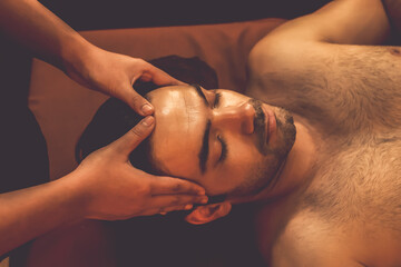 Naklejka premium Caucasian man enjoying relaxing anti-stress head massage and pampering facial beauty skin recreation leisure in warm candle lighting ambient salon spa in luxury resort or hotel. Quiescent