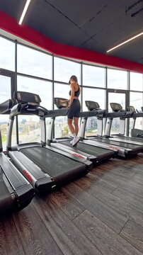Sofia, Bulgaria - 11.10.2025. A young woman in sportswear walks to a treadmill, adjusts the settings, and starts running in a modern gym with large panoramic windows and a city view. Vertical video.